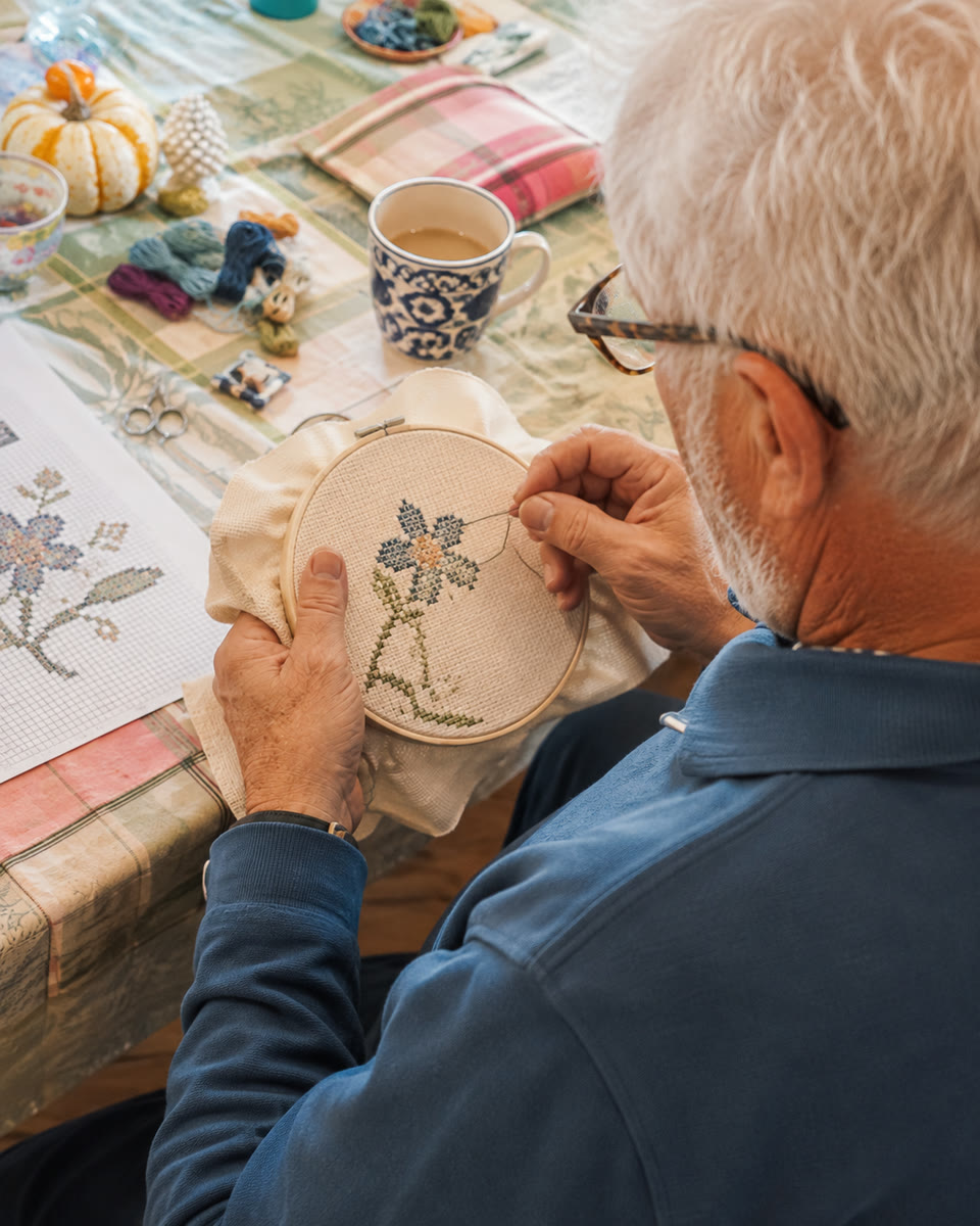 A stitcher working a flower in an embroidery hoop, threads and a coffee cup on the table beside him