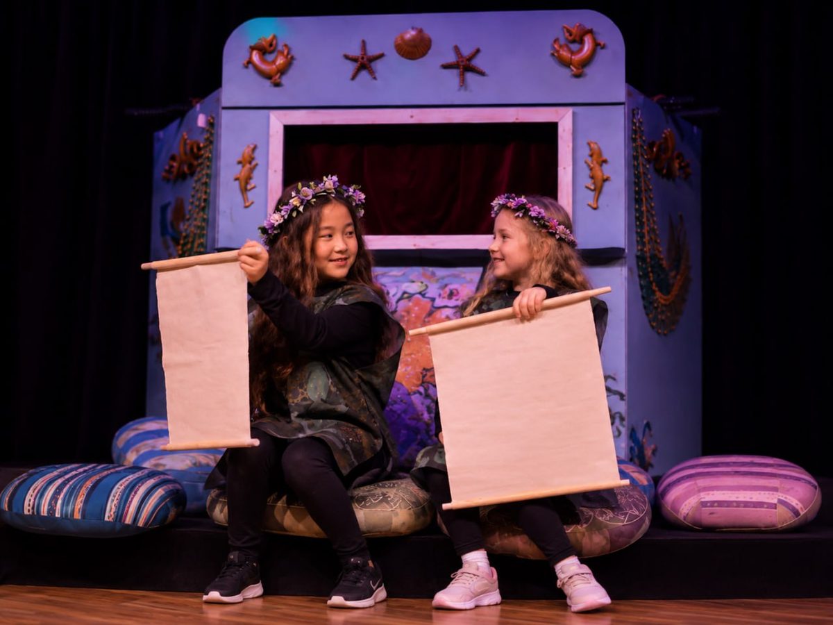 Two young performers on the Black Box stage, holding scrolls and laughing at each other