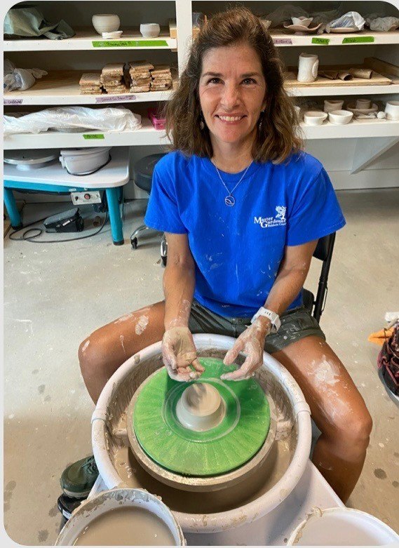 A potter at the wheel with the clay centered and the studio shelves behind.
