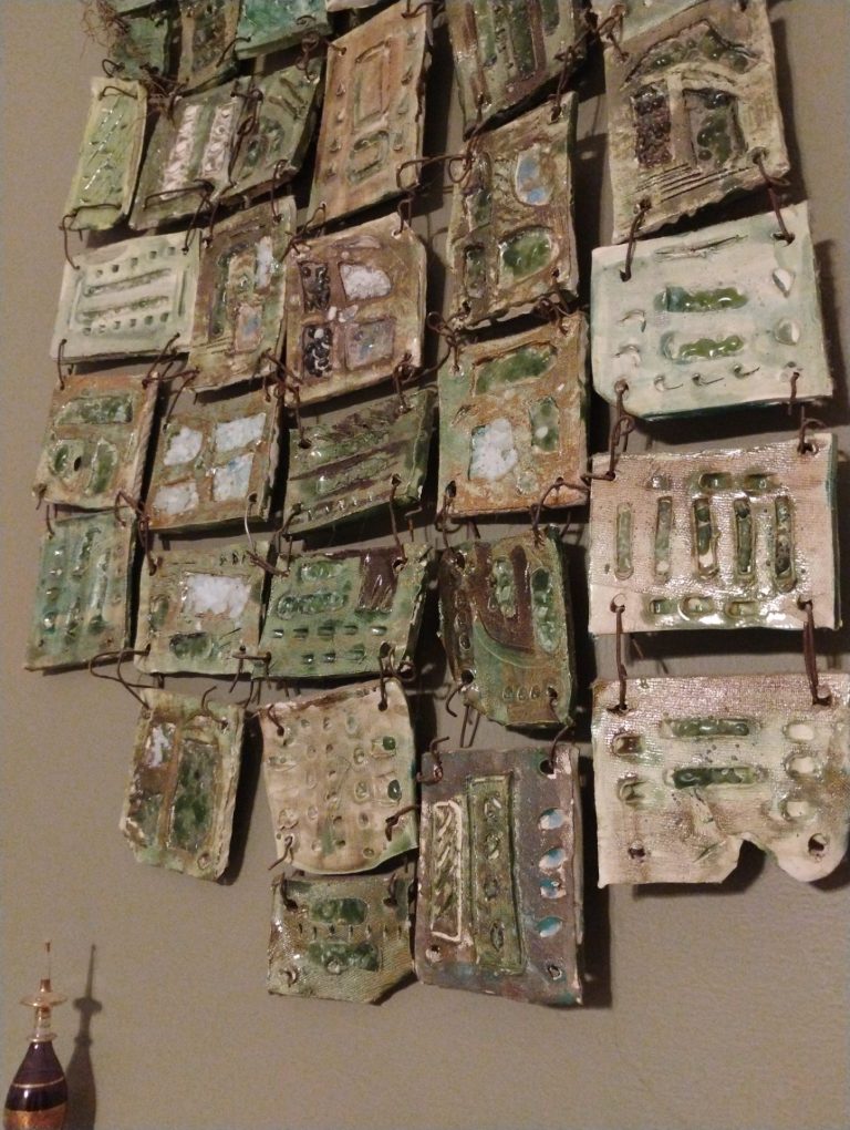 Dozens of small glazed clay tiles in greens and creams, wired together into a hanging quilt.