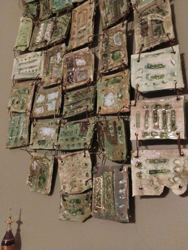 Dozens of small glazed clay tiles in greens and creams, wired together into a hanging quilt.