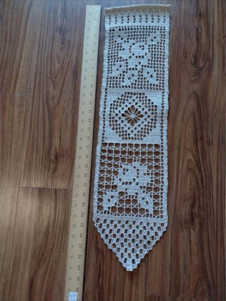 A filet crochet table runner in cream thread, laid on a wood floor beside a yardstick.