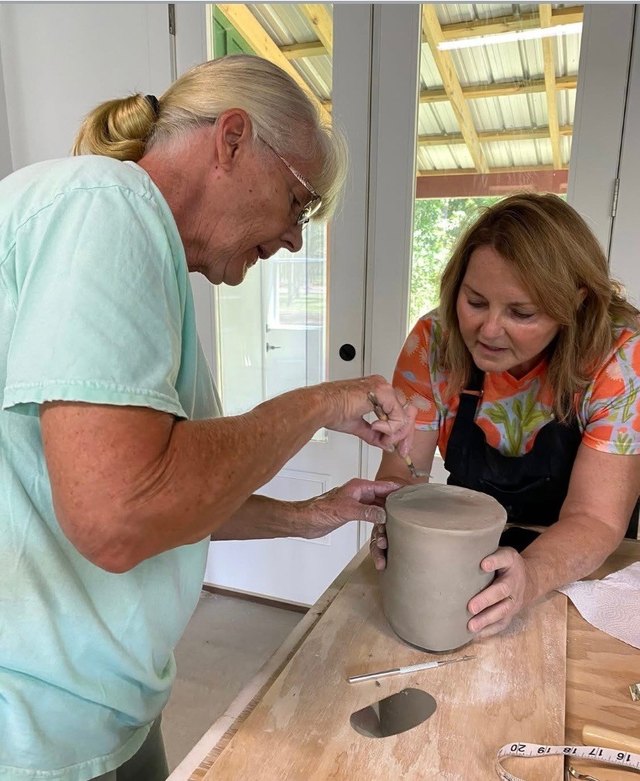 Two potters shaping a tall pot together at the studio table.