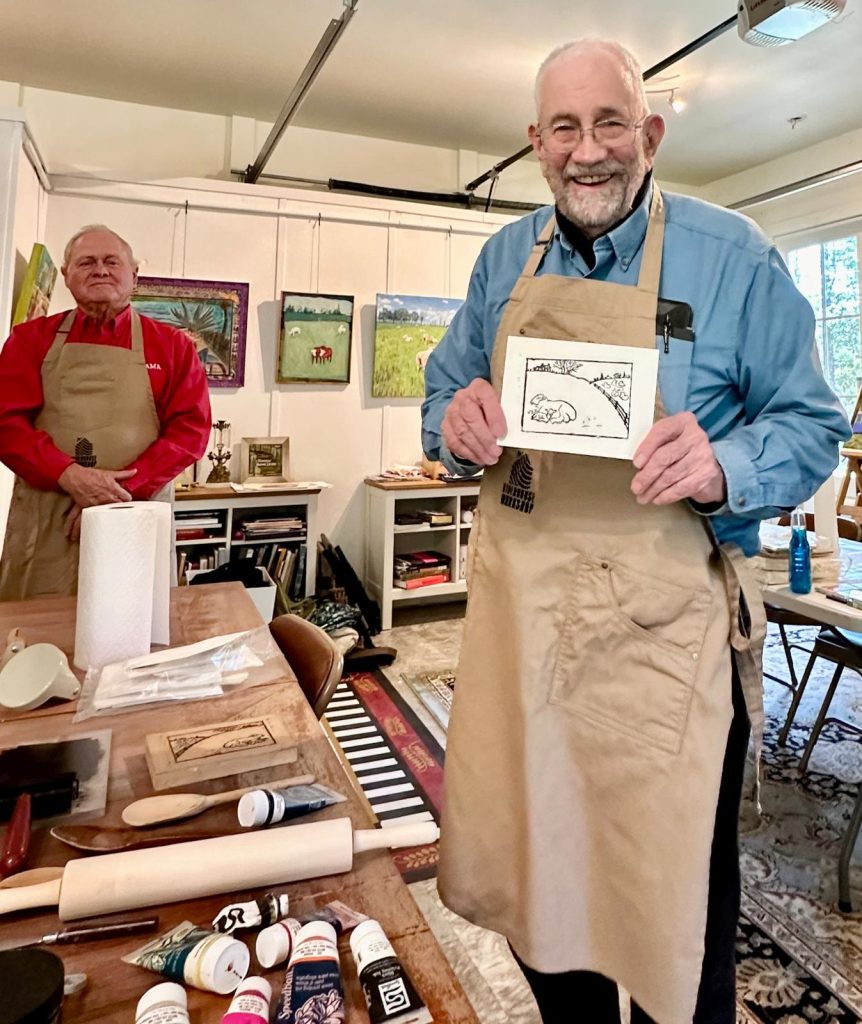 Two artists in aprons in a printmaking studio, one holding a finished print.