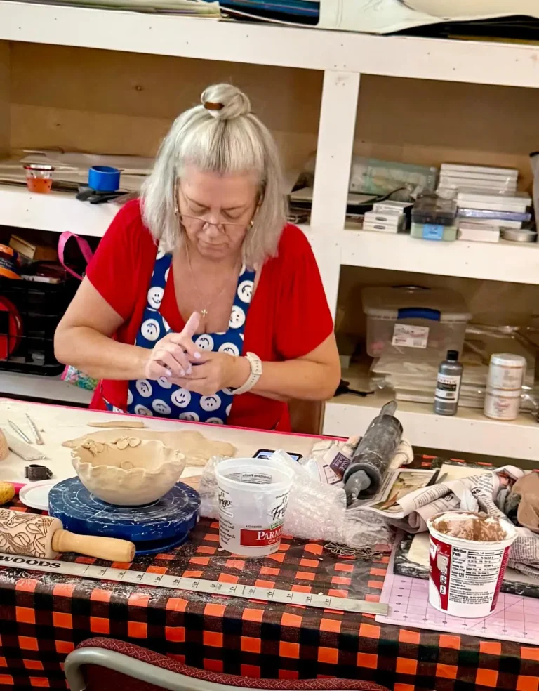 A potter hand-builds a clay bowl at a cluttered studio table