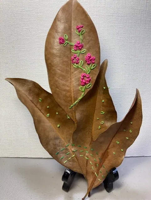 A dried leaf embroidered with small pink flowers, displayed upright