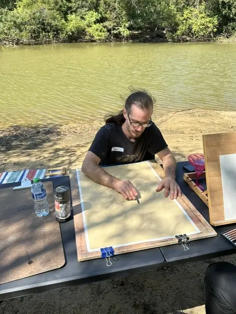 A man prepares a drawing board at a riverside picnic table