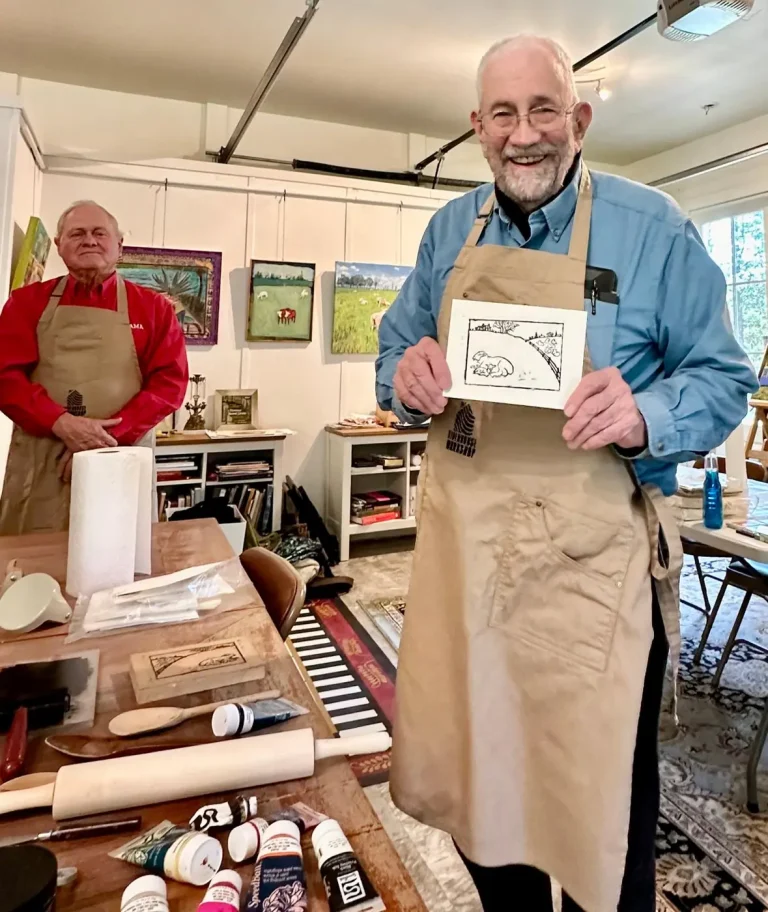 Two men in aprons in the print studio, one holding a landscape print